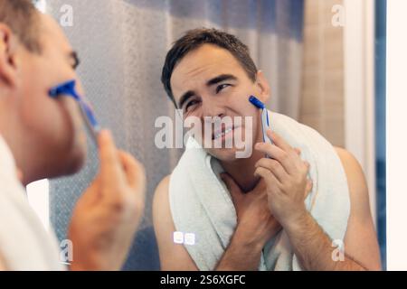 Ein Mann rasiert sein Gesicht ohne Creme oder Schaum und erlebt Schmerzen und Leiden. Trauriger Mann rasiert sich morgens vor einem Spiegel. Ungesunde Rasur von Stockfoto
