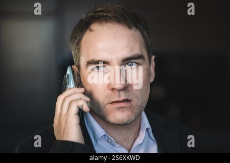 Ein hübscher Geschäftsmann mit einem verächtlichen Blick spricht nachts auf dem Handy vor dunklem Hintergrund. Nahporträt eines kaukasischen Mannes mit grauem stubb Stockfoto