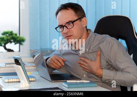 Büroleiter mit Herzinfarkt. Junger Mann, der die Hand auf schmerzhafter Brust am Arbeitsplatz hält. Medizinisches Konzept. Stockfoto