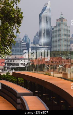 BANGKOK, THAILAND - 26. Februar 2024: Blick auf Bangkoks moderne Wolkenkratzer, die sich in einer Glasbarriere mit gebogenen Holzsitzen im widerspiegeln Stockfoto
