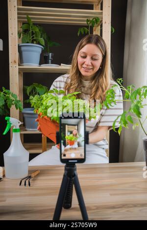 Eine junge Frau filmt ein Video-Tutorial über die Gartenarbeit im Haus, in dem Basilikumsämlinge in einem Topf gezeigt werden, während sie ein Smartphone auf einem Stativ für optimale Aufnahmen positioniert Stockfoto
