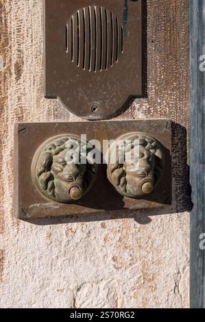 Antikes Bronze-Löwenkopf-Türklingeln auf einer hellbeigen Steinmauer. Eine alte Gegensprechanlage ist ebenfalls sichtbar. Stockfoto