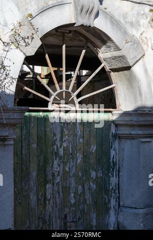 Verwitterte, bogenförmige Steintür mit einer verrottenden grünen Holztür und einem verzierten, rostigen Metallgitter. Ein Blick in die Vergangenheit. Stockfoto