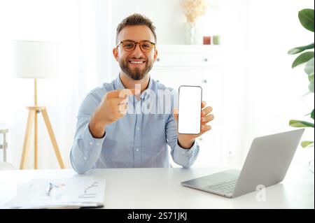Positiver, erfolgreicher Geschäftsmann, der am Schreibtisch im Büro sitzt und ein Smartphone mit leerem Modellbildschirm zeigt, mit dem Finger darauf zeigt, leeren Platz für Präsentationen oder Werbung Stockfoto