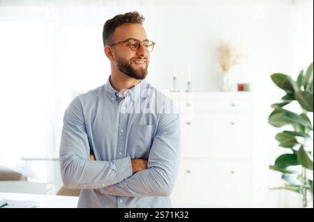 Selbstbewusster erfolgreicher kaukasischer oder arabischer Geschäftsmann mit Brille, in einem blauen Hemd gekleidet, firmenchef mit überkreuzten Armen im Büro stehend, lächelnd und zur Seite schauend Stockfoto