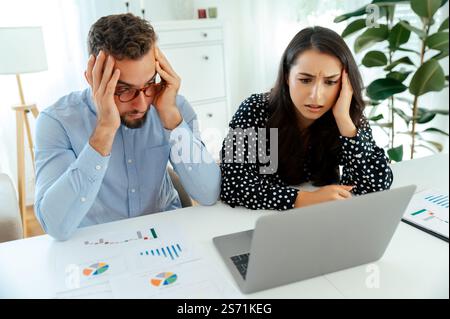 Gestresste Kollegen, die Daten und Diagramme auf einem Laptop analysieren und Blätter an einem Schreibtisch ausdrucken, arbeiten zusammen, um ein Problem zu lösen, während sie in einem modernen Büro sitzen, erleben Verwirrung und Enttäuschung Stockfoto