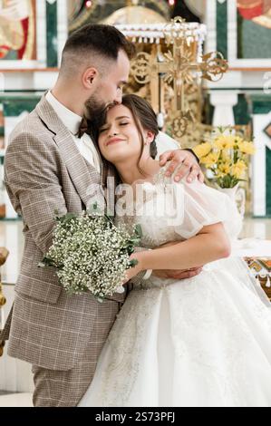 Ein frisch verheiratetes Paar teilt einen intimen Moment in einer Kirche. Der Arm des Bräutigams ist liebevoll um seine Braut, die ein schönes weißes Kleid trägt. Stockfoto