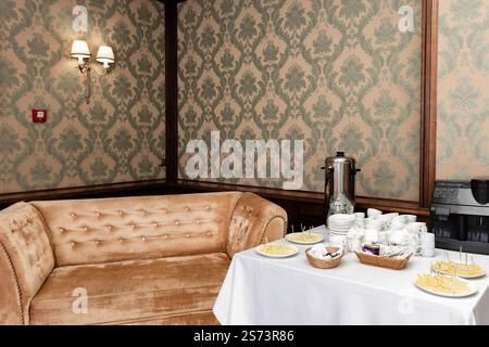 In einem Zimmer mit gemusterter Tapete befindet sich eine weiche Samtcouch. Auf einem Tisch werden Kaffee, Tee und kleine Snacks serviert, was eine Pause oder Wartezeit andeutet Stockfoto