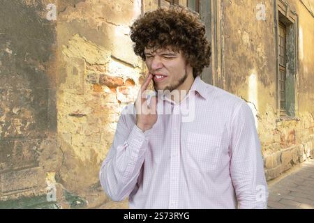 Hübscher Mann mit lockigem Haar, der seine Wange mit der rechten Hand berührt. Leiden an Zahnschmerzen. Alte schäbige Gebäudewand im Hintergrund. Stockfoto