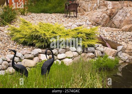 Erhöhte Felsengrenze mit Juniperus - Wacholdersträucher und schwarze Reiher Vogelskulpturen neben dem Teich im Vorgarten Zen Garten im Sommer, Quebec, Cana Stockfoto