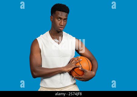 Junger afroamerikanischer Basketballspieler, der Ball hält und auf blauem Hintergrund selbstbewusst aussieht Stockfoto