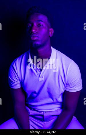 Porträt eines jungen schwarzen Mannes, der weißes Poloshirt und helle Hosen trägt und unter blauem Licht posiert Stockfoto