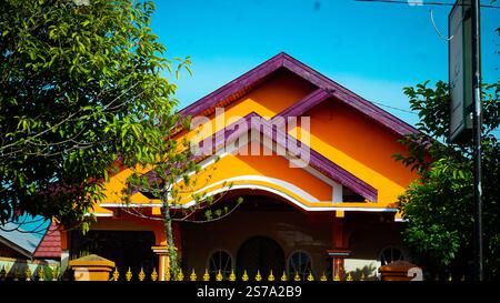 Ein lebhaftes oranges Haus mit violetten Dachakzenten, umgeben von üppigen grünen Bäumen unter einem klaren blauen Himmel Stockfoto