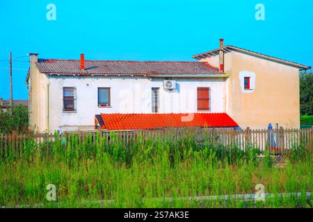 Rustikales Haus mit gekacheltem Dach und Gitterfenstern, umgeben von bewachsener Vegetation und einem Holzzaun Stockfoto