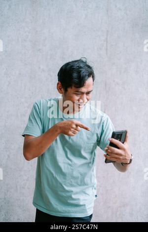 Asiatischer Mann, der aufgeregt auf sein Smartphone vor grauem Hintergrund zeigt. Stockfoto