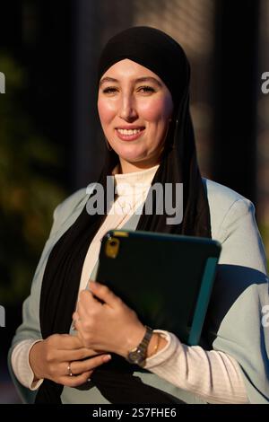 Lächelnde muslimische Geschäftsfrau, die Tablette hält und Hijab trägt Stockfoto