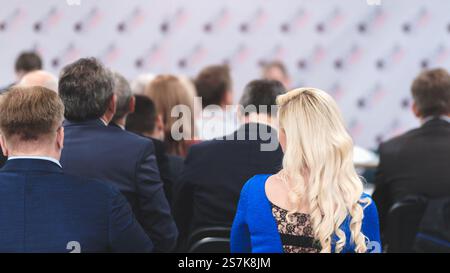 Eine Frau mit blonden Haaren sitzt in einem blauen Kleid in einem Raum voller Menschen. Das Zimmer ist mit viel weiß und rot dekoriert Stockfoto