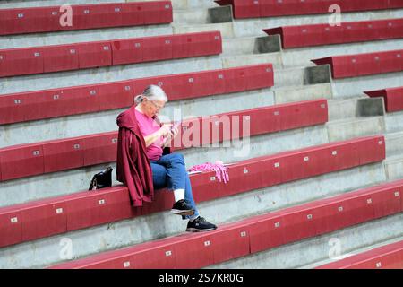 Eine ältere Frau, die allein in einer leeren Sportarena sitzt und auf ihrem Handy SMS schreibt oder surft, abgelenkt vom Basketballspiel auf der Arena. Stockfoto