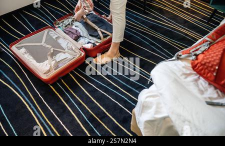 Eine Person, die Gegenstände in einem roten Koffer auf einem gestreiften Teppichboden organisiert und sich darauf vorbereitet, in einem Hotelzimmer mit gemütlicher Einrichtung und lebhaftem Dekor zu packen. Stockfoto