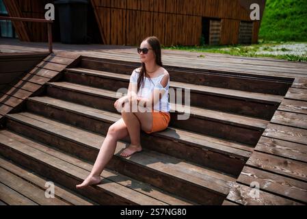 Eine barfüßige Russin in ihren 40ern sitzt im Sommer auf Holztreppen, trägt orange Shorts und eine weiße Bluse. Sie genießt das sonnige Wetter mit einem Schnaps Stockfoto