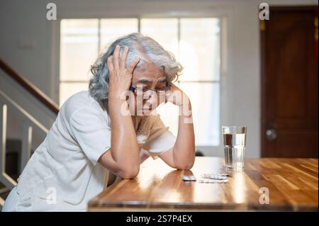 Eine traurige, frustrierte asiatische Seniorin sitzt an einem Esstisch in der Küche mit ihren Medikamenten auf dem Tisch und erlebt einen Angstanfall nach hinten Stockfoto