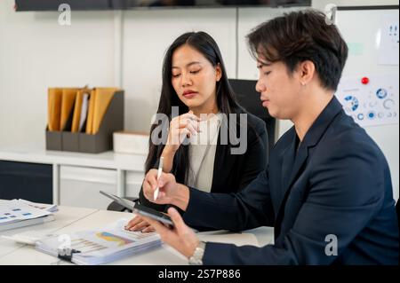 Ein professioneller und kluger asiatischer Geschäftsmann erklärt seiner Kollegin während eines Meetings im Büro die Arbeit auf seinem Tablet. Zwei fokussierte Busine Stockfoto