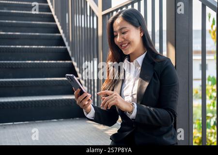 Eine schöne, glückliche asiatische Geschäftsfrau sitzt auf der Treppe, benutzt ihr Smartphone und plaudert während einer Pause mit ihren Freunden. Stockfoto
