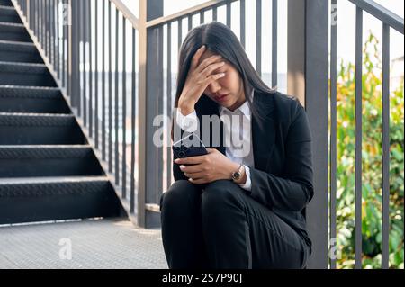 Eine gestresste, überforderte asiatische Geschäftsfrau sitzt auf der Treppe, bedeckt ihr Gesicht und weint, nachdem sie gefeuert wurde oder einen schlechten Arbeitstag hatte. Stockfoto