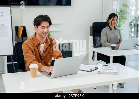 Ein positiver, gutaussehender asiatischer Geschäftsmann arbeitet neben seinen Kollegen im Büro an seinem Laptop. Geschäftsleute, Firmenjob, Marketingagentur Stockfoto