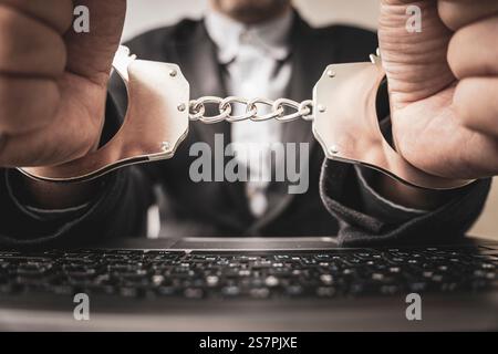 Ein Mann in Handschellen sitzt an seinem Arbeitsplatz mit einem Computer und Geld in Dollar. Das Konzept der Bestechung und Kriminalität im Cyber-Bereich. Stockfoto