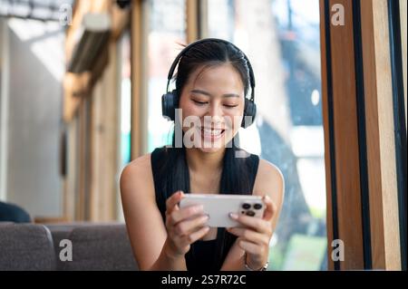 Eine fröhliche asiatische Frau mit Kopfhörern spielt gern ein mobiles Spiel oder schaut sich ein Video im Internet auf ihrem Smartphone an, während sie sich in einem Café entspannt. Stockfoto