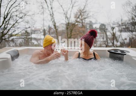 Seniorenpaar baden im Wannen-Whirlpool während des kalten Wintertages und toasten mit Champagner. Romantischer Wellness-Kurzurlaub mit Spa am Valentinstag-Wochenende. Stockfoto