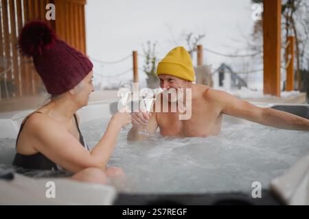 Seniorenpaar baden im Wannen-Whirlpool während des kalten Wintertages und toasten mit Champagner. Romantischer Wellness-Kurzurlaub mit Spa am Valentinstag-Wochenende. Stockfoto