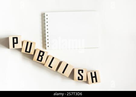 Ein weißes Notizbuch auf weißem Hintergrund. Das Wort „PUBLIZIEREN“ steht auf Holzblöcken Stockfoto