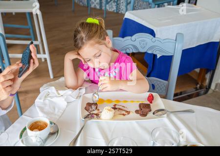 Vier Jahre altes blondes Mädchen, das süßes Dessert isst: Schwarze Schokolade, Vanilleeis, Erdbeeren und Mangosauce. Mutter mit Handy, sitzend Stockfoto