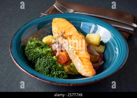 Gegrilltes Lachsfilet mit gedämpftem Brokkoli und geröstetem Gemüse auf blauem Keramikteller mit Gabel und Messer und brauner Serviette auf dunklem Hintergrund Stockfoto