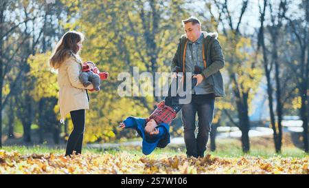Glückliche Familie, die einen sonnigen Herbsttag in einem Park genießt, Vater hält seinen Sohn kopfüber, während Mutter das Baby hält, umgeben von bunten gefallenen Blättern Stockfoto
