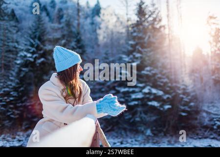 Die stilvolle junge Frau genießt den Blick auf den schneebedeckten Wald mit weißem, warmem Pelzmantel, gestricktem blauem Hut und Fäustlingen. Wintermode Stockfoto