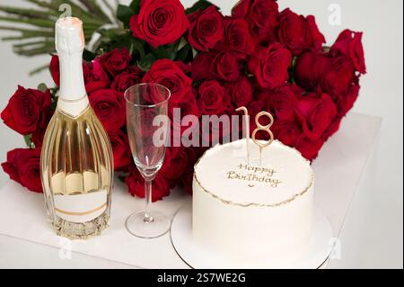 Ein festlicher Tisch bietet einen köstlichen Kuchen, eine Flasche Sekt, ein festliches Glas und einen atemberaubenden Strauß roter Rosen. Perfekt für ein 18. B Stockfoto