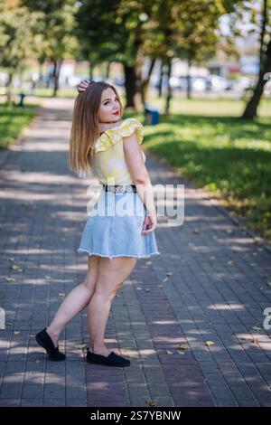Stilvolle Frau, die selbstbewusst durch den sonnendurchfluteten Park schreitet, fließende Haare mit Fingern, die Eleganz und moderne Raffinesse verkörpern Stockfoto