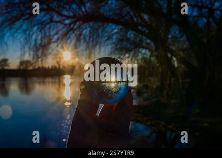Eine Person hält eine Glaskugel vor einem Gewässer. Die Sonne untergeht und strahlt ein warmes Licht auf die Szene. Die Reflexion der Sonne auf dem W Stockfoto