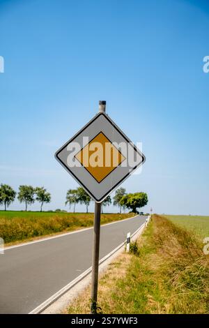 Ein gelb-weißes Schild befindet sich auf einer Straße in einem ländlichen Gebiet. Das Schild ist eine rautenförmige Form und wird auf einem Stab platziert. Der Himmel ist blau und es gibt Bäume im Stockfoto