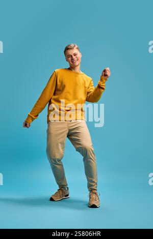 Dynamisches Ganzkörperfoto eines jungen Mannes, stilvoll gekleidet, tanzend, mit den Händen energetisch winkend vor blauem Studiohintergrund. Stockfoto