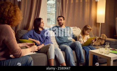 Familienberatung mit Psychologin, Mutter und Vater sitzen auf der Couch und äußern Probleme, und das Kind konzentriert sich auf das Zeichnen. Stockfoto