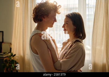 Seitenansicht eines wunderschönen jungen, kaukasischen Paares, das sich umarmt, während es im Wohnzimmer tanzt, erleuchtet von goldenen Sonnenstrahlen Stockfoto