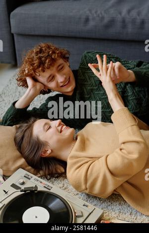 Vertikale Hochwinkelaufnahme einer kaukasischen Freundin und eines Freundes, die auf einem flauschigen Teppich liegen, lächelnd und spielend mit den Händen, während Musik vor dem Hintergrund spielt Stockfoto