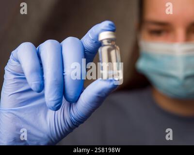 Medizinischer Facharzt untersucht klare Flüssigkeitsröhrchen, trägt blaue Latexhandschuhe und chirurgische Maske im klinischen Umfeld Stockfoto