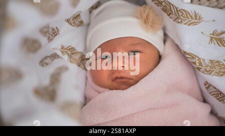 Das Neugeborene trägt einen weißen Hut mit einer kleinen Blumendekoration, in eine rosa Decke gewickelt und liegt in einer Krippe mit Federmustern Stockfoto