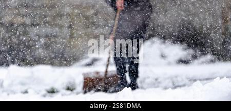 Eine Person fegt Schnee mit einer Schaufel. Der Schnee fällt und die Person trägt einen schwarzen Mantel Stockfoto