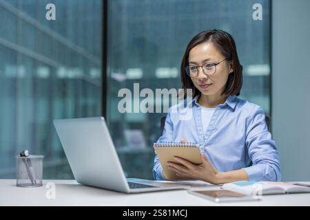 Seriöse und fokussierte asiatische Frau, die Online-Webinare und externe Meetings hört. Frau mitten im Büro, die mit einem Laptop arbeitet und Daten in ein Notebook schreibt. Stockfoto